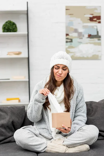 Больная молодая женщина в теплой одежде берет бумажную салфетку из коробки, сидя на диване — стоковое фото