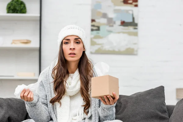 Хвора молода жінка в теплому одязі сидить на дивані вдома і тримає коробку з паперових серветок — стокове фото