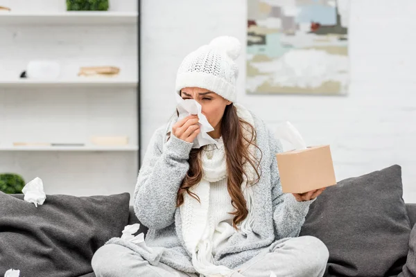 Jeune femme malade en vêtements chauds assis sur un canapé désordonné et éternuant avec des serviettes en papier à la maison — Photo de stock