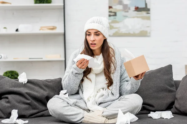 Хвора молода жінка в теплому одязі сидить на безладному дивані і чхає паперовими серветками вдома — стокове фото