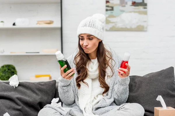 Jeune femme malade en vêtements chauds au choix entre des bouteilles rouges et vertes de liquides gargarisants à la maison — Photo de stock