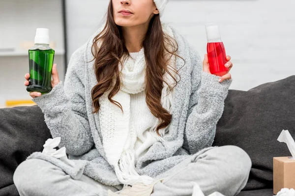 Plan recadré de jeune femme malade en vêtements chauds au choix entre des bouteilles rouges et vertes de liquides gargarisants à la maison — Photo de stock