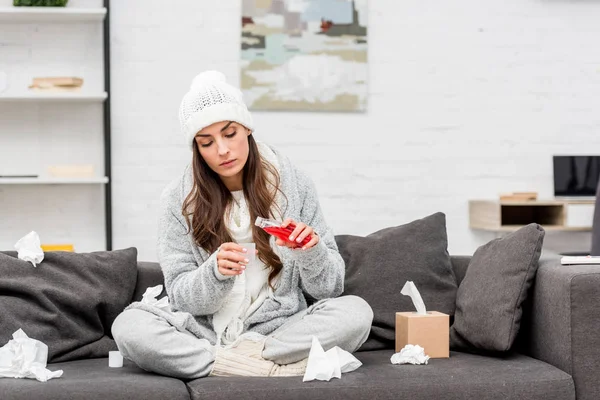 Kranke junge Frau in warmer Kleidung schüttet roten Hustensaft aus, während sie zu Hause auf der Couch sitzt — Stockfoto