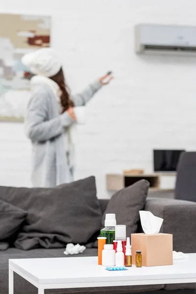 Femme malade en vêtements chauds pointant vers climatiseur avec télécommande avec divers médicaments debout sur la table au premier plan — Photo de stock