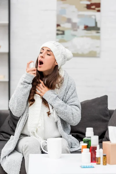 Хвора молода жінка в теплому одязі обприскує горло, сидячи на дивані вдома — стокове фото