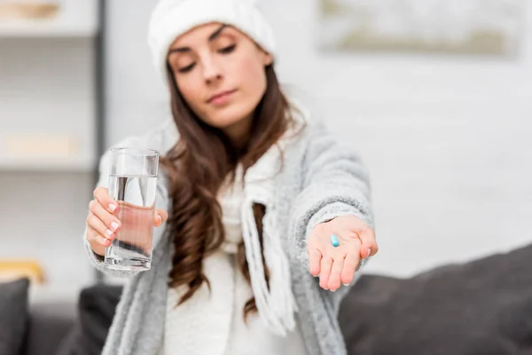 Sick young woman in warm clothes holding glass of water and showing pill in hand at home — Stock Photo