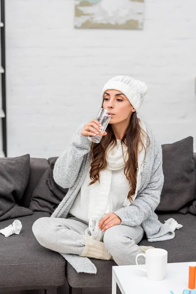 Kranke junge Frau in warmer Kleidung trinkt Wasser auf der heimischen Couch — Stockfoto