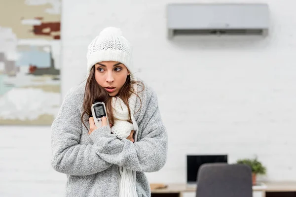 Congélation jeune femme en vêtements chauds tenant la télécommande avec climatiseur sur fond — Photo de stock