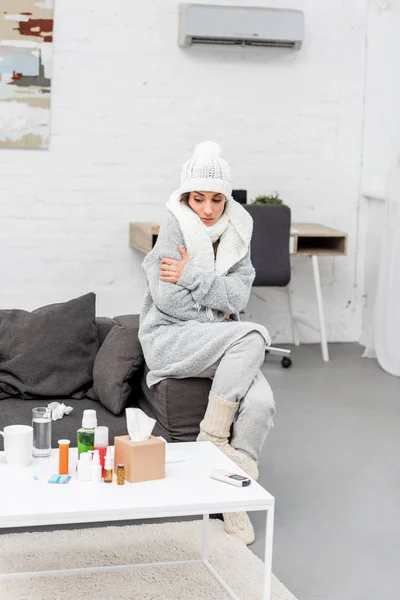 Sick young woman in warm clothes sitting on couch and looking at medicines on table — Stock Photo