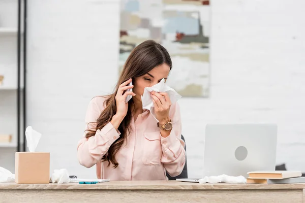 Sick young businesswoman talking by phone and sneezing at workplace — Stock Photo