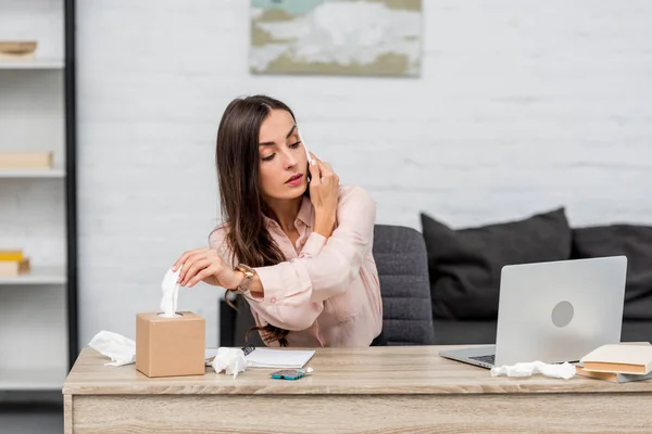 Sick young businesswoman talking by phone and taking paper napkin out of box at workplace — Stock Photo
