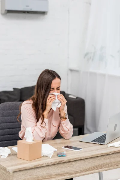 Kranke junge Geschäftsfrau pustet Nase mit Papierserviette am Arbeitsplatz — Stockfoto