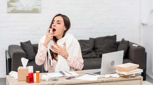 Sick young businesswoman spraying throat at workplace — Stock Photo