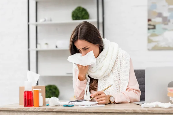 Sick young businesswoman sneezing with paper napkin at workplace — Stock Photo