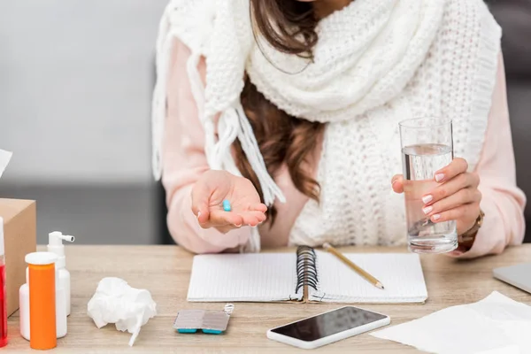 Plan recadré de jeune femme malade en écharpe tenant verre d'eau et pilule sur le lieu de travail — Photo de stock