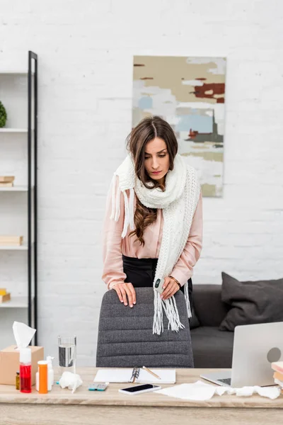 Хвора молода жінка в шарфі, що стоїть біля робочого місця в офісі — стокове фото