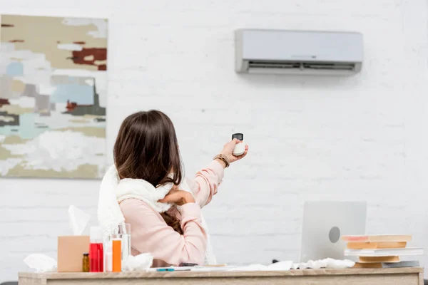 Sick young woman sitting at workplace and pointing at air conditioner with remote control — Stock Photo
