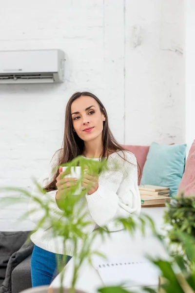 Belle jeune femme tenant une tasse de café tout en s'appuyant sur le rebord de la fenêtre — Photo de stock