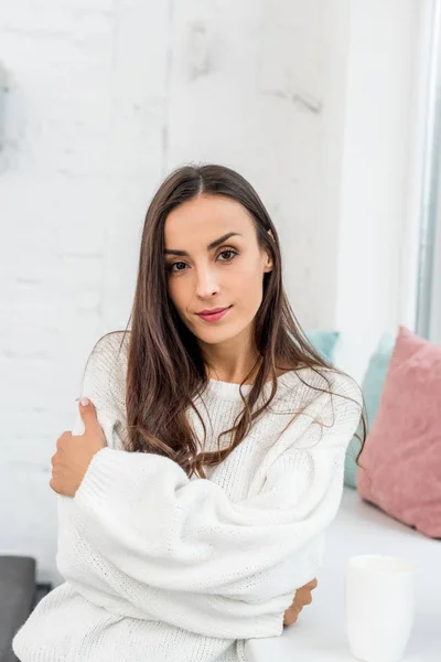 Beautiful young woman in white sweater looking at camera near window — Stock Photo