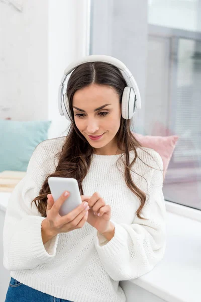Smiling young woman listening music with smartphone and wireless headphones ner window — Stock Photo