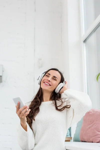 Mujer joven sonriente escuchando música con smartphone y auriculares inalámbricos en casa - foto de stock