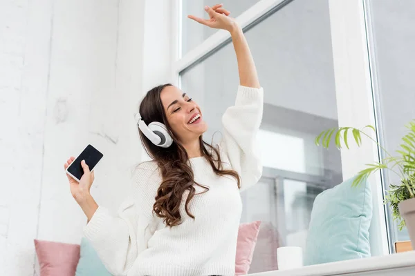 Vista inferior de la joven feliz escuchando música con smartphone y auriculares inalámbricos en casa - foto de stock