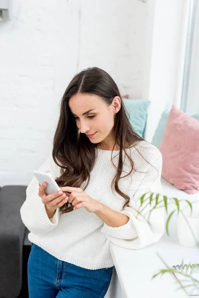 Belle jeune femme en utilisant smartphone près du rebord de la fenêtre à la maison — Photo de stock