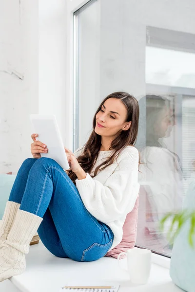 Mujer joven feliz usando la tableta en el alféizar de la ventana con los cojines en el país - foto de stock