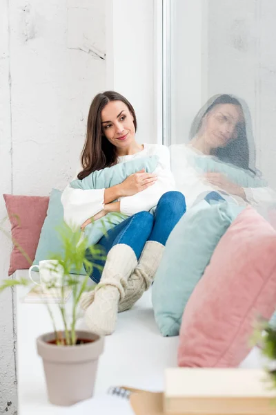 Beautiful young woman relaxing on windowsill with cushions at home — Stock Photo