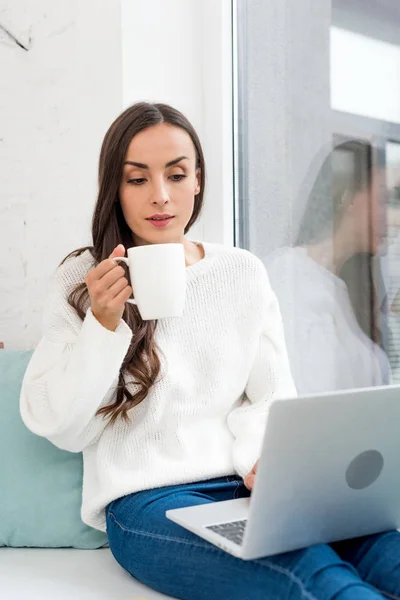 Hermosa joven freelancer beber café y trabajar con el ordenador portátil en el alféizar de la ventana en casa - foto de stock