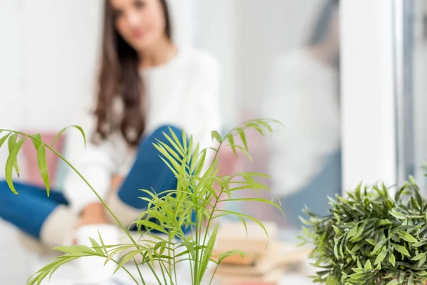 Selektiver Fokus der schönen jungen Frau, die zu Hause auf der Fensterbank sitzt, mit Zimmerpflanzen im Vordergrund — Stockfoto