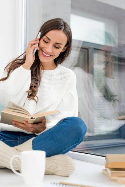 Hermosa joven leyendo libro y hablando por teléfono en el alféizar de la ventana en casa - foto de stock