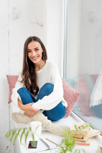 Hermosa joven sentada en el alféizar de la ventana en casa y mirando a la cámara - foto de stock