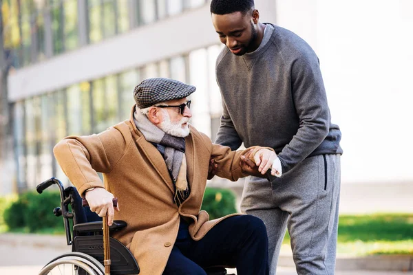 Молодой африканский американец помогает пожилому инвалиду встать с инвалидного кресла на улице — стоковое фото