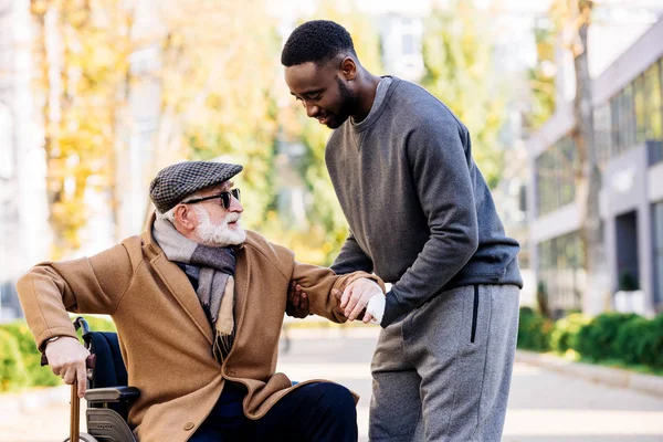 Young african american cuidador helping senior disabled man to get up from wheelchair on street — Stock Photo