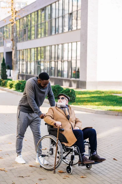 Senior disabled man in wheelchair with walking stick and african american cuidador talking on street — Stock Photo