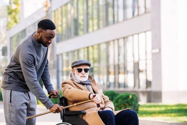 Afro-américain donnant bâton de marche à un homme handicapé âgé en fauteuil roulant dans la rue — Photo de stock