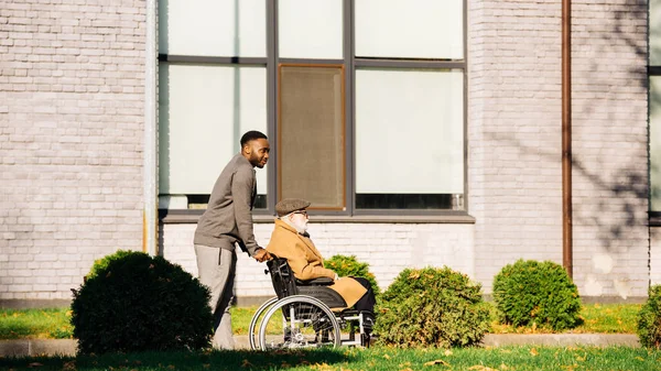 Senior handicapé en fauteuil roulant et homme afro-américain ayant promenade matinale dans la rue — Photo de stock