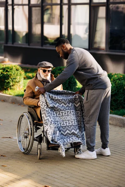 African american nurse covering senior disabled man in wheelchair with plaid on street — Stock Photo