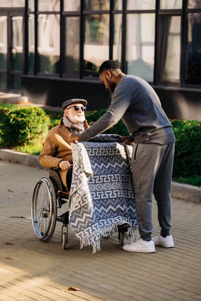 Joven africano americano cuidador cubriendo sénior discapacitado en silla de ruedas con cuadros en calle - foto de stock