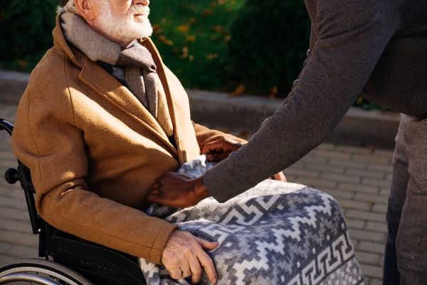 Schnappschuss eines Afroamerikaners, der behinderten Senior im Rollstuhl mit Plaid auf der Straße bedeckt — Stockfoto