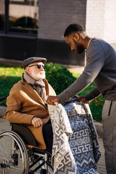 Afro-américain homme couvrant aîné handicapé en fauteuil roulant avec plaid sur la rue — Photo de stock