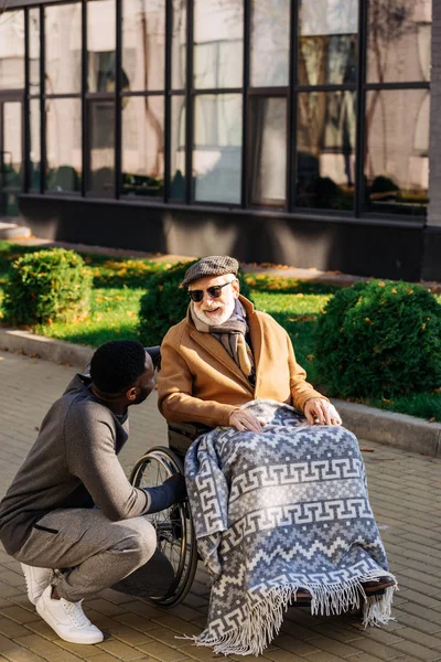 Homem deficiente sênior feliz em cadeira de rodas e enfermeira americana africana conversando na rua — Fotografia de Stock