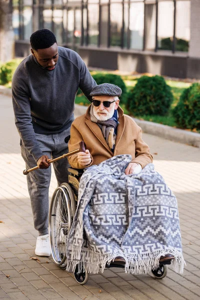 Africano americano hombre dando caminar palo a mayor discapacitado hombre en silla de ruedas en calle - foto de stock