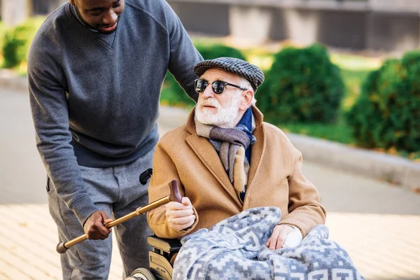 Young african american man giving walking stick to senior disabled man in wheelchair on street — Stock Photo