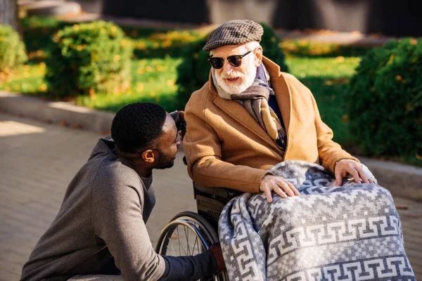 Anziano disabile uomo in sedia a rotelle e africano infermiera americana in chat per strada — Foto stock