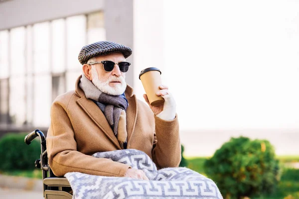 Anciano discapacitado en silla de ruedas sosteniendo taza de papel de café en la calle - foto de stock