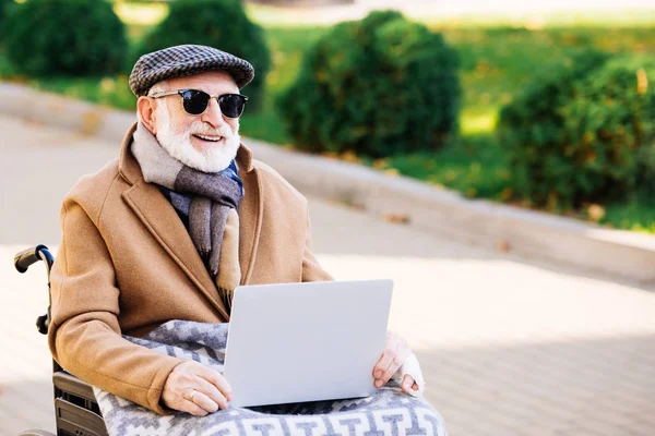 Felice uomo anziano disabile in sedia a rotelle utilizzando il computer portatile sulla strada — Foto stock