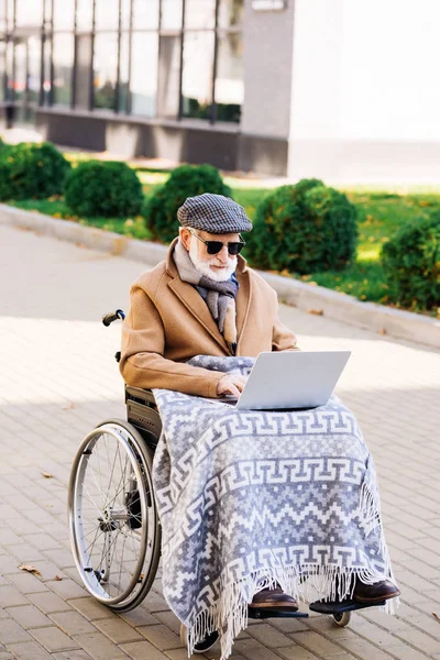 Senior disabled man in wheelchair with plaid working with laptop on street — Stock Photo