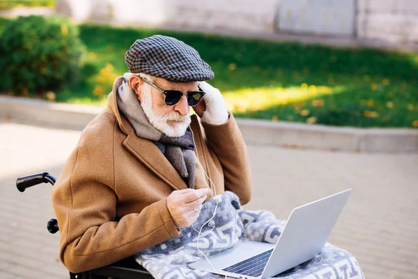 Schöner älterer behinderter Mann im Rollstuhl mit Laptop und Kopfhörer auf der Straße — Stockfoto